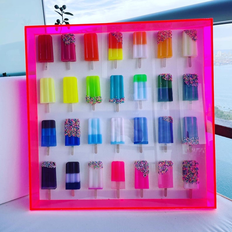 Puede incluir: Un marco rosa ne&oacute;n contiene una colecci&oacute;n de 24 piezas de arte de resina con forma de paleta. Cada paleta est&aacute; decorada con chispas de colores y tiene un color y un patr&oacute;n diferente.