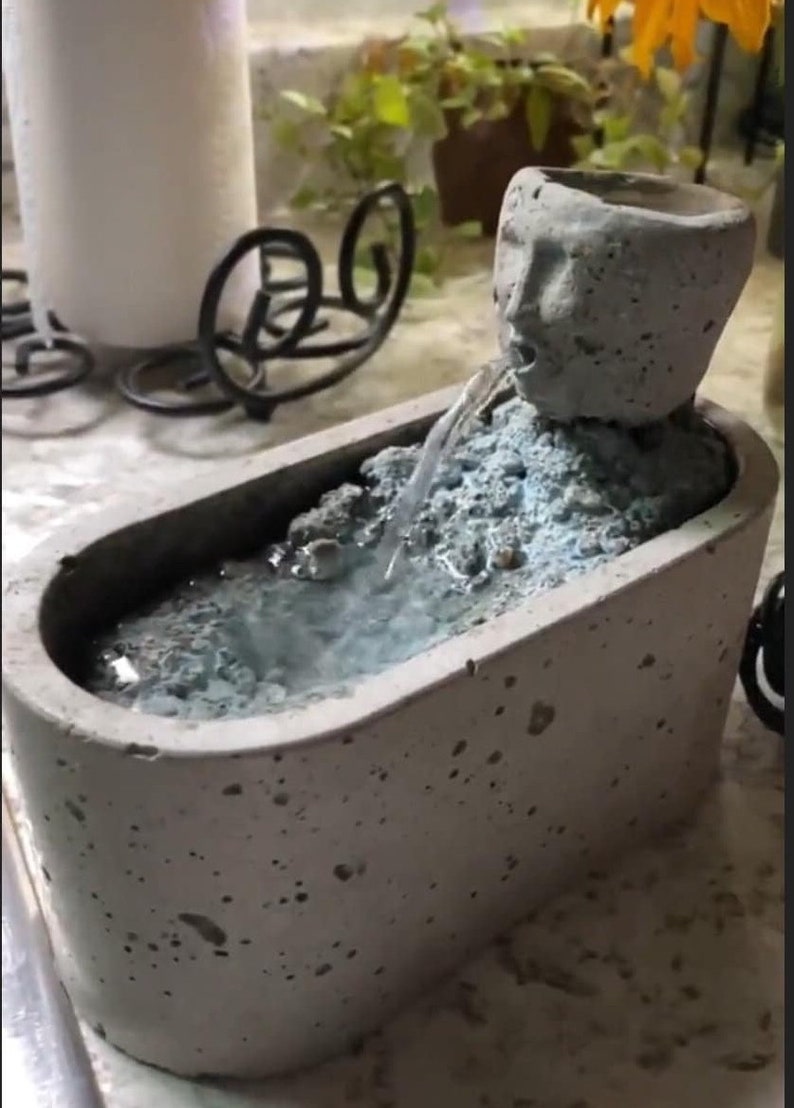 K&ouml;nnte beinhalten: Ein kleiner Betonbrunnen mit einem auf der Oberseite modellierten Gesicht. Der Brunnen ist badewannenf&ouml;rmig und hat einen kleinen Wasserstrahl, der aus dem Gesicht in die Wanne flie&szlig;t.