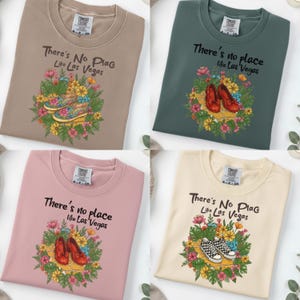 May include: Four crew neck sweatshirts in various colours: beige, green, pink, and cream. Each shirt displays the text "There's No Place Like Las Vegas" above a floral design with shoes. The shoes vary in style and colour.