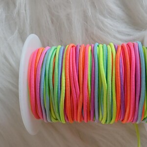 May include: A spool of colorful elastic cord in shades of pink, orange, yellow, green, blue, and purple. The cord is tightly wound on a white plastic spool, ideal for crafting and jewelry making.