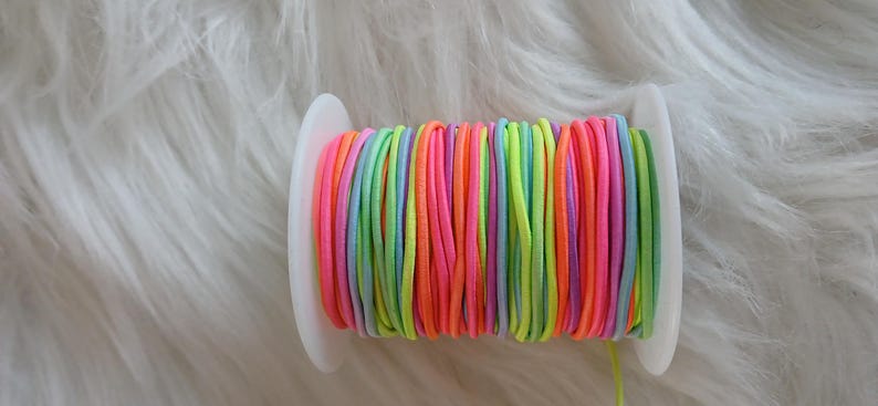 May include: A spool of colorful elastic cord in shades of pink, orange, yellow, green, blue, and purple. The cord is tightly wound on a white plastic spool, ideal for crafting and jewelry making.