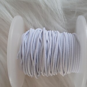 May include: A spool of light gray elastic cord. The cord is tightly wound around a white plastic spool. The background is a soft, white, furry texture.