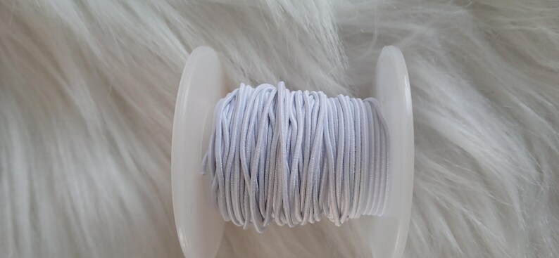 May include: A spool of light gray elastic cord. The cord is tightly wound around a white plastic spool. The background is a soft, white, furry texture.