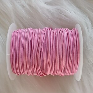 May include: A spool of pink elastic cord, tightly wound on a white plastic bobbin. The cord is a vibrant shade of pink, and the bobbin is set against a fluffy, white background. This is a craft supply, ideal for sewing and jewelry making.