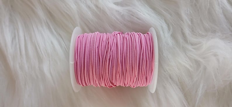May include: A spool of pink elastic cord, tightly wound on a white plastic bobbin. The cord is a vibrant shade of pink, and the bobbin is set against a fluffy, white background. This is a craft supply, ideal for sewing and jewelry making.