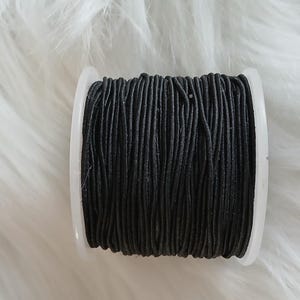 May include: A spool of black elastic cord, tightly wound on a white plastic bobbin. The cord appears to be made of many thin strands. The background is a soft, white, fluffy material.