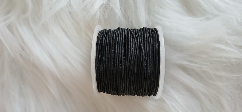 May include: A spool of black elastic cord, tightly wound on a white plastic bobbin. The cord appears to be made of many thin strands. The background is a soft, white, fluffy material.