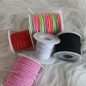 May include: Spools of colorful elastic cord in red, pink, white, black, and a rainbow of colors. The cords are wound on white plastic spools, perfect for crafting and DIY projects. The image is set against a fluffy white background.