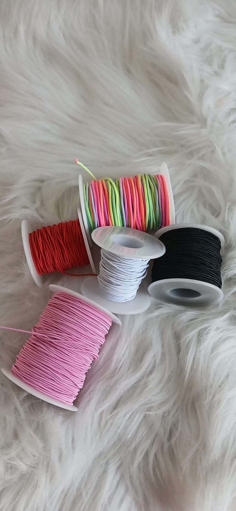 May include: Spools of colorful elastic cord in red, pink, white, black, and a rainbow of colors. The cords are wound on white plastic spools, perfect for crafting and DIY projects. The image is set against a fluffy white background.