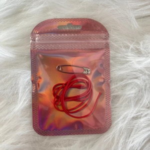 May include: A small, iridescent pink pouch containing a silver safety pin and a length of red cord. The pouch is sealed and has a small hole at the top. The background is a soft, white, textured surface.