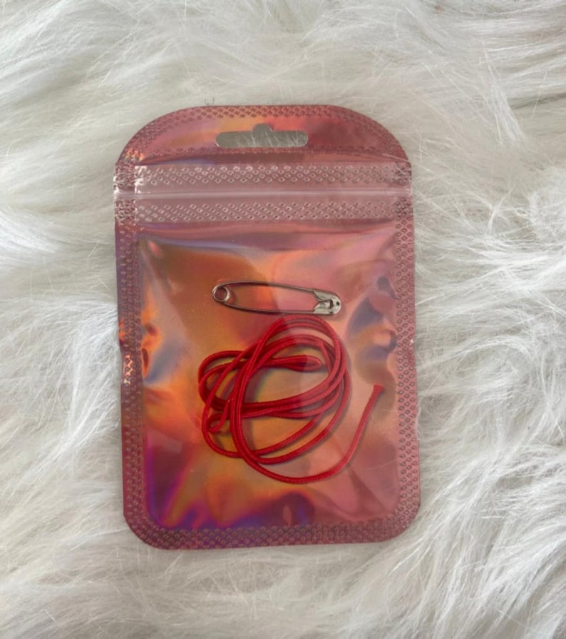 May include: A small, iridescent pink pouch containing a silver safety pin and a length of red cord. The pouch is sealed and has a small hole at the top. The background is a soft, white, textured surface.