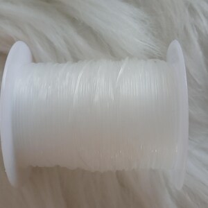 May include: A spool of clear, elastic beading cord. The cord is tightly wound around a white plastic spool. The image is set against a soft, white, textured background, suggesting a craft or jewelry-making theme.