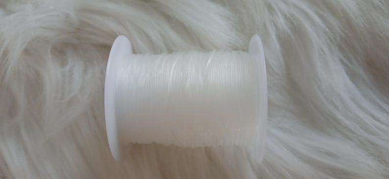 May include: A spool of clear, elastic beading cord. The cord is tightly wound around a white plastic spool. The image is set against a soft, white, textured background, suggesting a craft or jewelry-making theme.