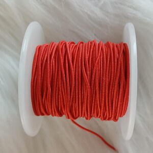 May include: A spool of bright red cord, tightly wound between two white plastic discs. The cord is a vibrant shade of red, and the spool is set against a soft, white, textured background.