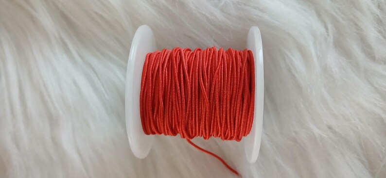 May include: A spool of bright red cord, tightly wound between two white plastic discs. The cord is a vibrant shade of red, and the spool is set against a soft, white, textured background.