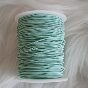 May include: A spool of mint green elastic cord. The cord is tightly wound around a white plastic spool. The background is a soft, white, fluffy material.