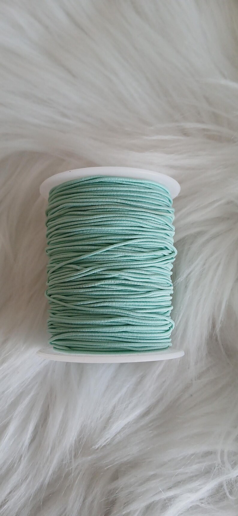 May include: A spool of mint green elastic cord. The cord is tightly wound around a white plastic spool. The background is a soft, white, fluffy material.