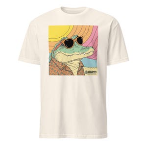 May include: Cream-colored t-shirt featuring a graphic of an alligator wearing sunglasses and a patterned shirt, set against a colorful sunset backdrop. The text "LIGIPRO relaxed coastal vibes" is also visible.