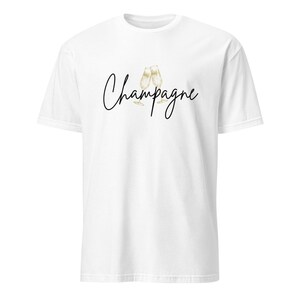 May include: White t-shirt with the word "Champagne" in a black script font, accompanied by a gold illustration of two champagne flutes. The shirt is a classic crew neck style.