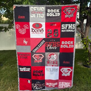 May include: A rectangular memory quilt constructed from various red, black, and gray fabric squares featuring printed text and logos including Spanish Fork High School, Dons, Cheer, Rock Solid, Hope Squad, and the number 28 with a gray fuzzy border.