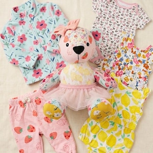 Pode incluir: Um urso de peluche com um laço rosa e saia de tule rosa, rodeado por um casaco de lã azul claro com flores, calças rosa com estampado de morangos, um body branco com flores, um top floral e um macacão amarelo com estampado de limões.