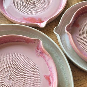Könnte beinhalten: Rosa Keramik-Reibeplatten mit einer spiralförmig strukturierten Oberfläche. Die Platten haben einen kleinen Ausguss an der Seite und sind mit einer glänzenden Oberfläche glasiert. Die Platten sind in verschiedenen Größen auf einer Holzoberfläche angeordnet.