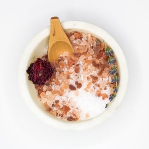 May include: A white ceramic bowl filled with pink Himalayan salt, white sea salt, dried lavender buds, and a dried rose. A small wooden spoon is resting on top of the salt.