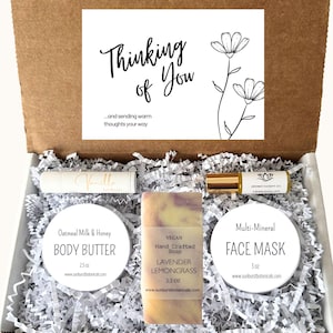 May include: A gift box with a white floral card that reads "Thinking of You." The box contains a lip balm, a bar of lavender lemongrass soap, a body butter, a face mask, and an aromatherapy oil.
