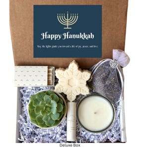 May include: A gift box filled with Hanukkah themed items, including a white snowflake-shaped soap, a small potted succulent plant, a white candle, a small bag of lavender, and a box of matches. The box is decorated with white tissue paper and a card that reads "Happy Hanukkah" with a menorah graphic. The card also includes the text "May the lights guide you toward a life of joy, peace, and love."