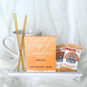 May include: A white mug with a white saucer, two yellow straws, a box of "Cup of Sunshine" herbal tea with golden turmeric and ginger, and two individually wrapped Rip Van Wafels with Dutch Caramel and Vanilla flavors.