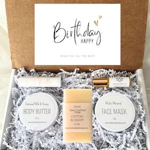 May include: A gift box filled with self-care products. The box contains a white jar of oatmeal milk and honey body butter, a white jar of multi-mineral face mask, a bar of vegan hand-crafted cotton blossom soap, a small tube of vanilla lip balm, and a small bottle of aromatherapy oil. The box is decorated with white shredded paper and a card that says "Birthday Happy Wish You All The Best".