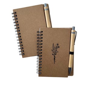 May include: Two spiral-bound notebooks with kraft paper covers. One notebook has a black floral design. Two bamboo pens are included. The notebooks are ideal for journaling, note-taking, or sketching.