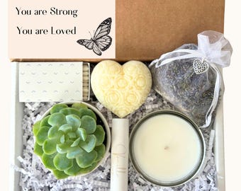 May include: A gift box filled with self-care items, including a succulent plant, a heart-shaped soap, a candle, a lip balm, and a small bag of lavender. The box also includes a card with the message "You are Amazing, You are Strong, You are Loved" and a butterfly illustration.