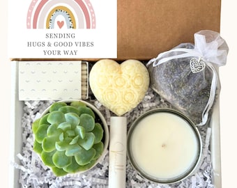 May include: A gift box filled with self-care items, including a succulent plant, a heart-shaped soap, a candle, a lip balm, and a lavender sachet. The box is decorated with white shredded paper and a card that reads "Sending hugs & good vibes your way".
