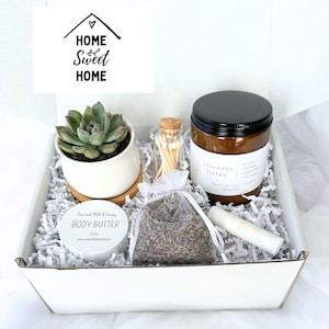 May include: A white gift box filled with a succulent in a white pot, a jar candle, body butter, a bag of lavender, matches, and a lip balm. The box has shredded paper and a "Home Sweet Home" graphic.