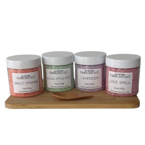 May include: Four clear jars of foaming bath salts with white lids, labeled Sweet Orange, Eucalyptus Mint, Lavender, and Love Spell. Each jar contains 5 oz / 142 g of bath salts. The jars sit on a wooden tray with a small wooden scoop.
