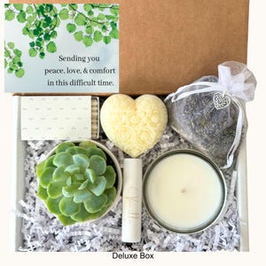 May include: A white gift box filled with items for self-care and relaxation. The box includes a small succulent plant, a heart-shaped soap, a candle, a tube of lip balm, a small bag of lavender, and matches. The box is labeled "Deluxe Box".