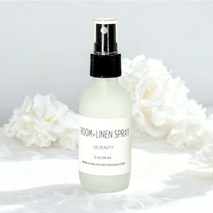 May include: A frosted glass bottle of "Room + Linen Spray" with a black spray nozzle. The label reads "Serenity" and "2 oz | 59 ml". The background features white flowers, creating a clean and calming aesthetic.