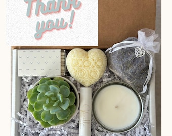 May include: A gift box containing a green succulent in a white pot, a heart-shaped soap with a rose pattern, a white lip balm labeled Vanilla, a candle in a glass jar, a bag of dried lavender, and a box of matches. A card reads Thank you!