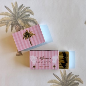 May include: A rectangular matchbox with a pink and white striped pattern featuring a palm tree illustration. The box is printed with the text Alyssa's PALM HOUSE Boca Raton and contains several wooden matches with dark brown tips.