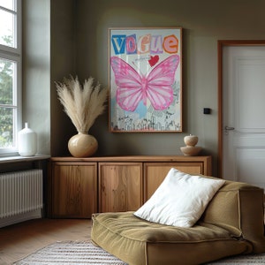May include: A wooden cabinet with three doors, a vase with dried pampas grass, a pink butterfly illustration with the word "Vogue" in colourful letters, and a brown ottoman with a white cushion.