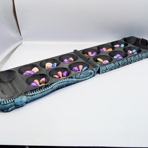May include: A black Mancala board game with colorful stones. The board is divided into two sections, each with six circular pits. The pits are filled with a variety of small, colorful stones. The board has a textured, decorative border.