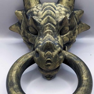 Peut inclure: Un heurtoir de porte en forme de tête de dragon de couleur dorée avec une poignée en forme d'anneau. Le visage du dragon est détaillé avec des écailles et une finition texturée.