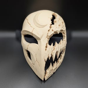 May include: A beige, dual-sided mask featuring a smooth, cracked surface with a crescent moon on the left side and a jagged, cratered, toothy texture on the right side. The mask has large, hollow eye openings and a pointed, tapered chin design.