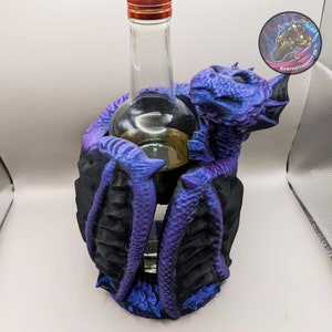 Puede incluir: Un porta botellas de dragón morado y negro con una botella de vidrio transparente en su interior. El dragón tiene un acabado texturizado e iridiscente.