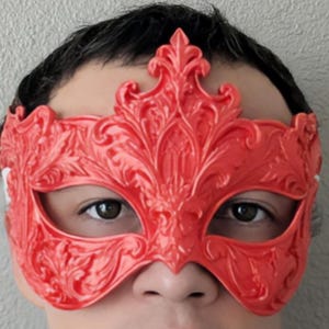 May include: A bright red masquerade mask featuring an intricate, raised filigree pattern with a prominent fleur-de-lis style crest at the center. The mask covers the upper face, including the eye area, and is secured with a white elastic strap.