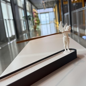 May include: A white cherub figurine incense holder featuring a small winged figure standing on a black rectangular base. The figurine holds a thin brown incense stick horizontally, extending outward from the cherub's hands over the black tray.