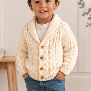 May include: A cream-colored cable knit cardigan with a shawl collar and four brown buttons, worn over a plain white t-shirt. The outfit is completed with medium-wash blue denim jeans and light gray sneakers with hook-and-loop closure straps.