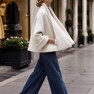 May include: A woman wearing a cream-colored cape-style jacket and dark blue wide-leg jeans walks on a city sidewalk. The jacket has a textured weave. She wears white flats. The background includes a storefront and the sign "Max Mara."