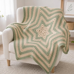 Op de afbeelding: Een gehaakte deken met een groot sterpatroon in afwisselende strepen van saliegroen en crèmekleurig garen. De deken is gedrapeerd over een witte fauteuil en toont een gestructureerd steekontwerp dat vanuit de centrale ster naar buiten straalt.